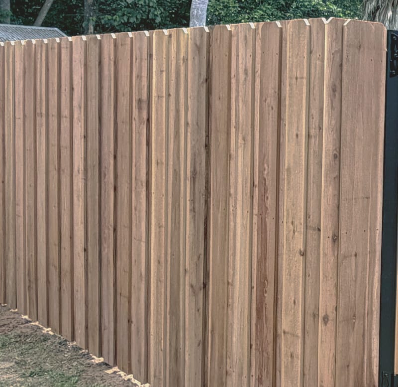A privacy fence of tight vertical boards with dog ear tops turning an outside corner, with a slim black steel post at the corner and trees behind.