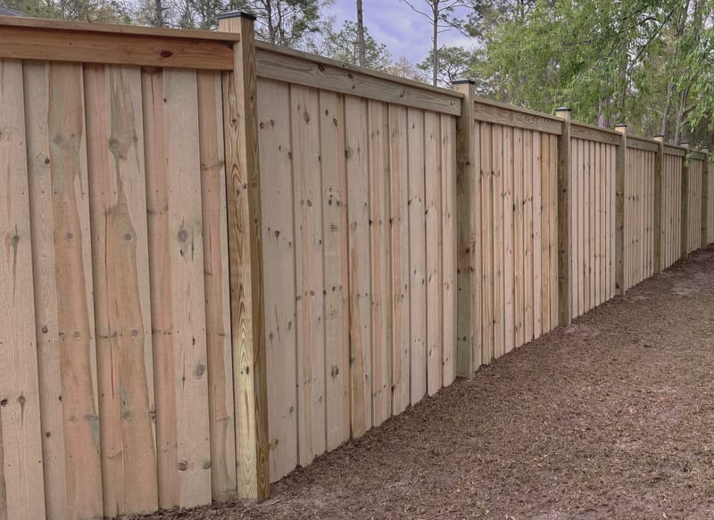 A wood privacy fence photographed along its length from a low angle, showing overlapping vertical boards, a flat capped top rail and black post caps stepping away into the distance.